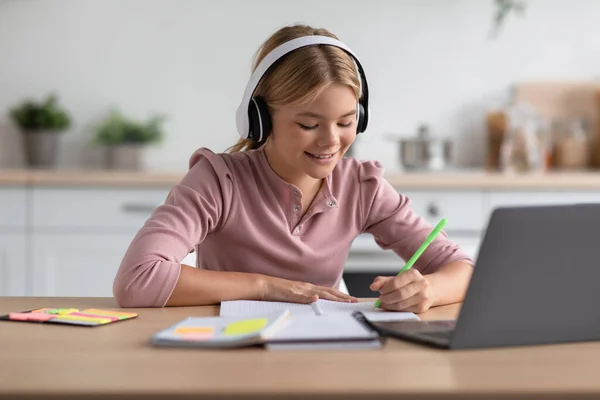 Blij Europese jongeren meisje blond leerling in hoofdtelefoon maakt notities, studeren thuis aan tafel — Stockfoto