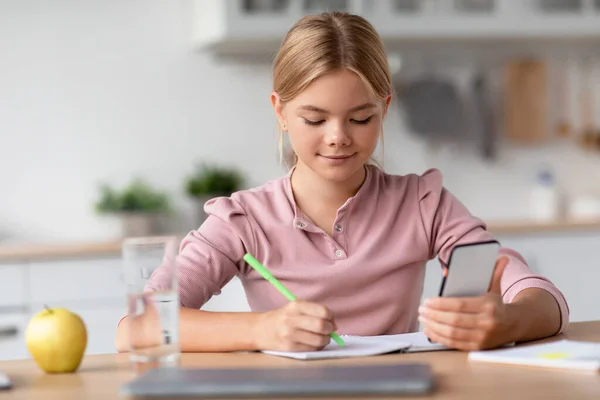 Vážná hezká blondýnka evropský teenager dívka dělá poznámky s smartphonem u stolu v minimalistická kuchyně — Stock fotografie
