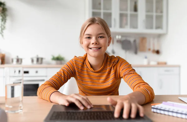 快乐的欧洲少女金发姑娘在舒适的厨房里用笔记本电脑在键盘上打字 — 图库照片