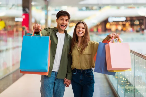 Cônjuges alegres mostrando sacos de compras para câmera em pé no shopping — Fotografia de Stock