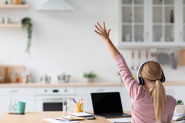 Caucasian teen girl blonde in headphones study at home, raises her hand for answer looks at pc with blank screen