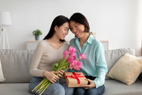Ung asiatisk kvinna med mogen mamma håller blommor och presentask, firar födelsedag eller mors dag tillsammans hemma — Stockfoto
