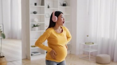 Neşeli Hamile Asyalı Kadın Kulaklıkla müzik dinliyor ve evde dans ediyor