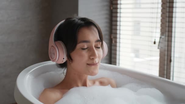 Женщина принимает ванну, слушая музыку в наушниках в ванной комнате — стоковое видео
