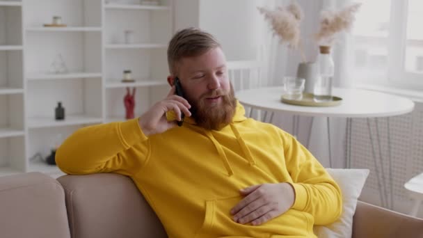 Szakállas fickó beszél mobilon ül a kanapén otthon — Stock videók