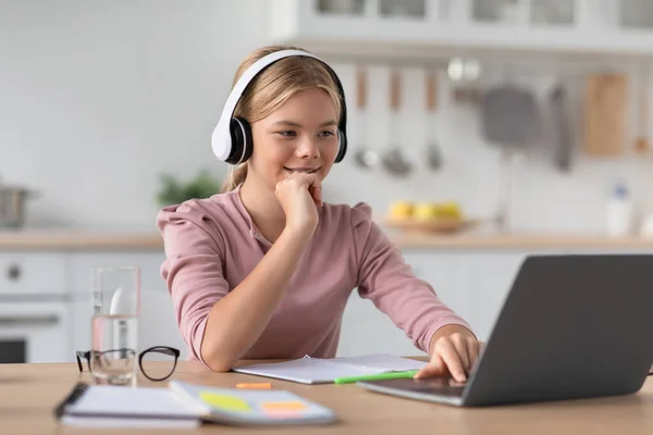Heureux heureux caucasien jeune fille blonde dans les écouteurs à table avec ordinateur portable dans l'intérieur de la cuisine, espace libre — Photo