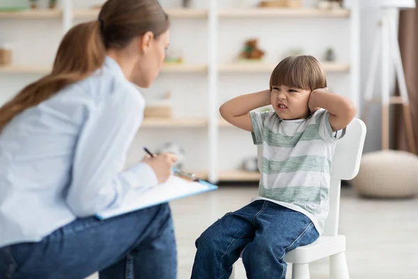Professioneller Psychotherapeut, der versucht, mit schwierigen Kindern zu sprechen, mürrischem kleinen Jungen, der Ohren verdeckt und die Stirn runzelt — Stockfoto