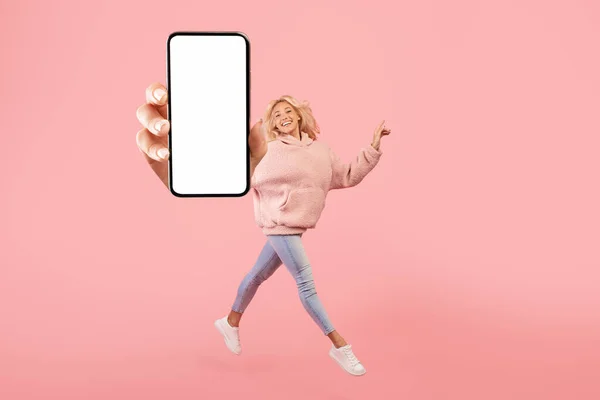 Glückliche junge Frau zeigt leeren Smartphone-Bildschirm, während sie über rosa Studiohintergrund springt, Collage, Attrappe — Stockfoto