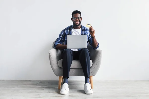 Красивий молодий чорний чоловік з кредитною карткою і ноутбуком сидить в кріслі, купуючи в Інтернеті, роблячи безконтактну оплату — стокове фото