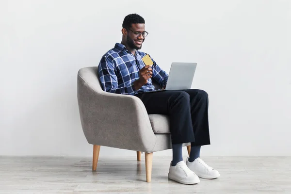 Positivo jovem negro segurando cartão de crédito, usando laptop em poltrona, compras na internet, fazendo pagamento sem contato — Fotografia de Stock