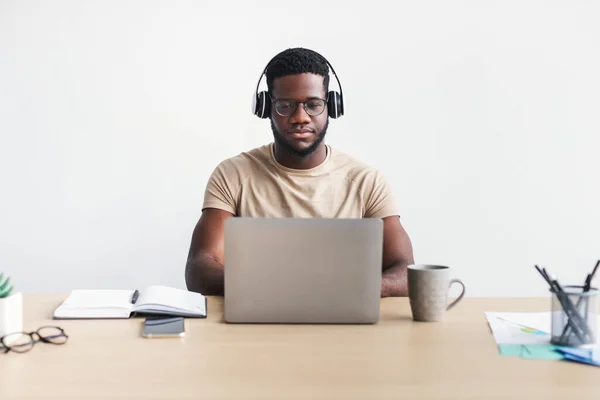 Фокусований молодий чорний хлопець в навушниках спілкується онлайн, маючи ділову зустріч на ноутбуці за столом — стокове фото