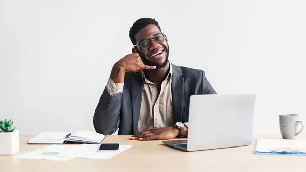 Θετικός μαύρος επιχειρηματίας με επίσημο κοστούμι που δείχνει χειρονομία τηλεφωνικής κλήσης στο τραπέζι με φορητό υπολογιστή σε λευκό φόντο στούντιο — Φωτογραφία Αρχείου