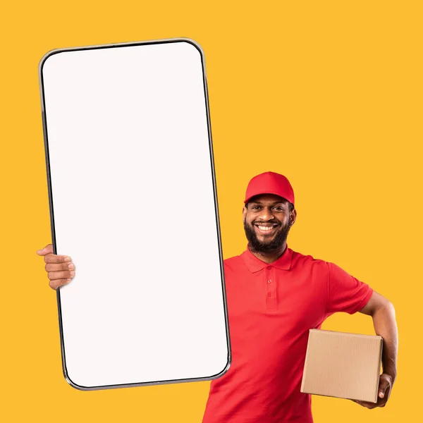 Uomo di consegna nero barbuto dimostrando cellulare con schermo bianco e contenente scatole di cartone, mockup — Foto Stock