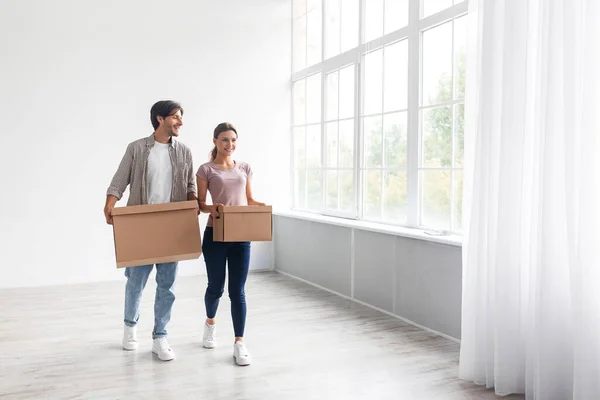 Satisfied european young man and lady in casual carry cardboard boxes in empty room interior with window — Stock Photo, Image