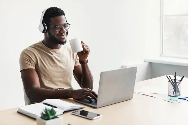 Позитивний молодий чорний хлопець в навушниках п'є каву, використовуючи ноутбук за столом в домашньому офісі, копіювати простір — стокове фото