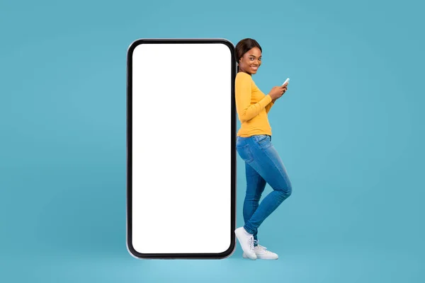 Cheerful Black Woman Holding Cellphone And Leaning At Big Blank Mobile Phone