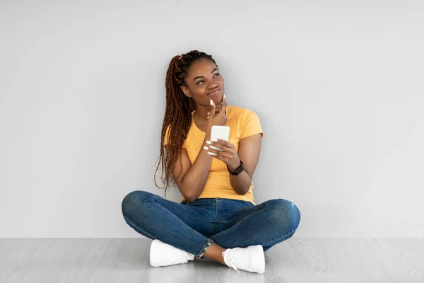Задумчивая молодая черная женщина, сидя скрестив ноги на полу, используя смартфон, думает о чем-то против серой стены — стоковое фото