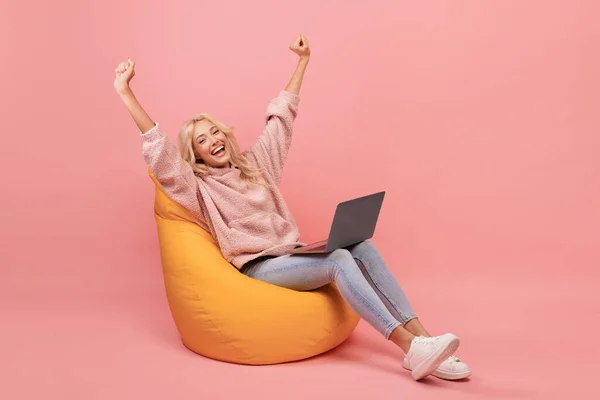 Overjoyed woman in casual sitting in beanbag chair with laptop and raising hands up, got dream job or great news