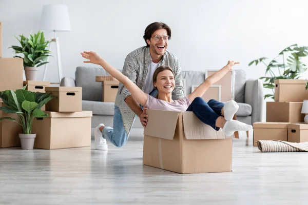 Zadowolony biały millenium facet popycha żonę w pudełku w pokoju wnętrza, zabawy razem cieszyć się przeprowadzką do nowego domu — Zdjęcie stockowe