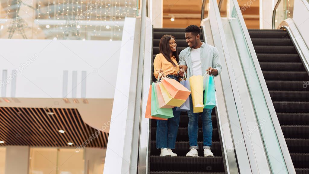 Happy black couple holding shopping bags going on escalator