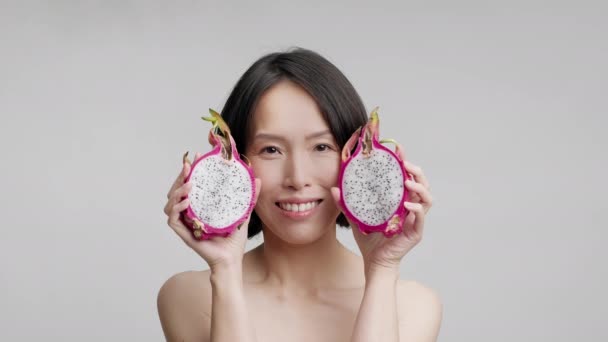 Mujer asiática madura sosteniendo frutas de dragón cerca de la cara, fondo gris — Vídeos de Stock