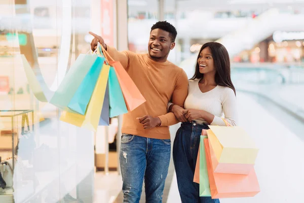 Cheerful black couple holding shopping bags pointing at window — Stock Photo, Image