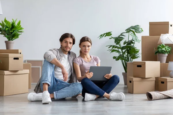 Serious europeo millennial dama y chico en casual mira tableta sentarse en el suelo con cajas de cartón —  Fotos de Stock