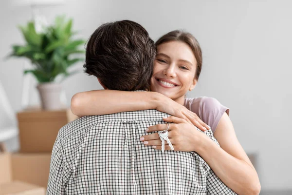 Feliz esposa milenaria europea abrazando marido y sosteniendo las llaves en la habitación con cajas de cartón —  Fotos de Stock