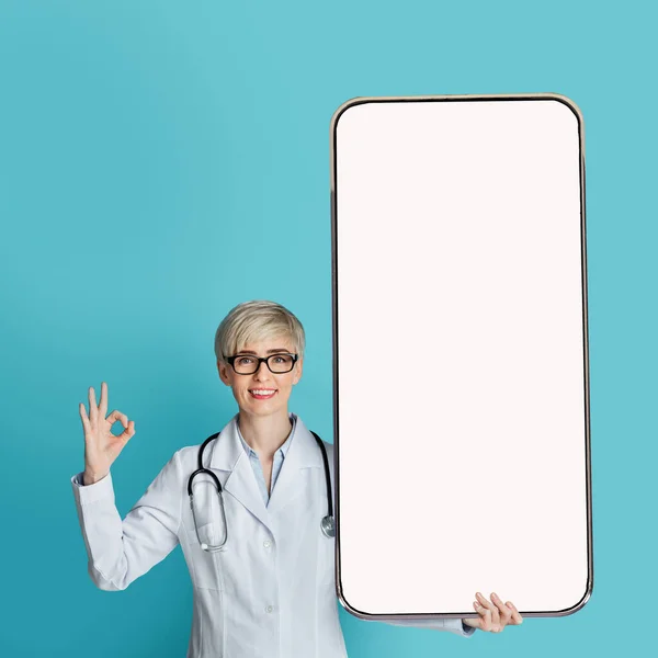 Medico donna che mostra grande smartphone con schermo bianco e gesticolando segno OK, in piedi sopra lo sfondo blu, mockup — Foto Stock