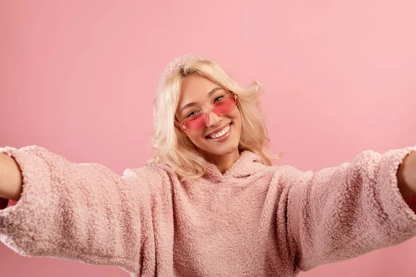 幸せなブロガーの女性は、自分自身を作ります,カラフルなメガネとパーカーを身に着けています,ポーズとカメラに笑顔,ピンクの背景 — ストック写真