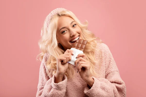 Fuskmåltid. Glada dam bita chokladkaka, äta skräpmat och titta på kameran, står över rosa bakgrund — Stockfoto