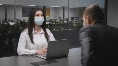 Covid-19 salgını sırasında iş toplantısı. Modern ofiste koruyucu yüz maskesi takan genç bir kadın müşteriyi selamlıyor.