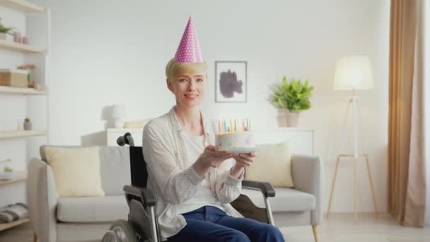 Mujer madura positiva con discapacidad con gorra de fiesta soplando velas en pastel de cumpleaños y sonriendo a la cámara, acercar — Vídeos de Stock