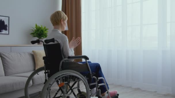 Kobieta w średnim wieku na wózku inwalidzkim patrząca na okno sama, rozgrzewająca ręce i odjeżdżająca w domu, powiększenie — Wideo stockowe