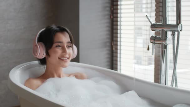 浴室でリラックスして入浴するヘッドフォンを身に着けている陽気な女性屋内 — ストック動画