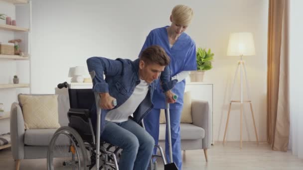 Udana rehabilitacja. Mężczyzna niepełnosprawny wstaje z wózka inwalidzkiego z pomocą pielęgniarki, opierając się na kulach — Wideo stockowe
