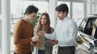 Satış temsilcisi genç mutlu müşterilere işaret veriyor. Araç galerisinde evrakları imzalayan adam.