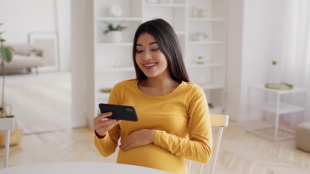 Feliz joven embarazada asiática mujer relajante con teléfono móvil en casa — Vídeos de Stock