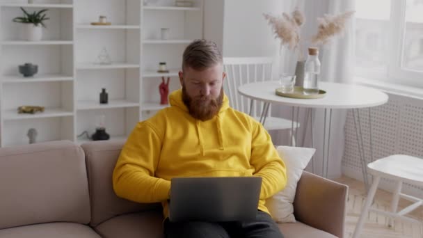 Man freelancer met behulp van laptop werken online zitten op de bank binnen — Stockvideo