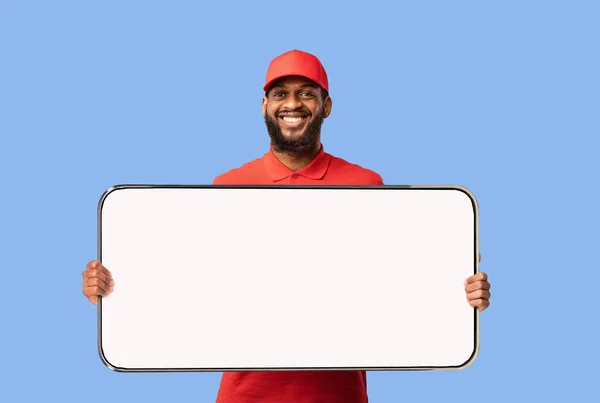 Corriere maschio afroamericano che mostra un grande cellulare con schermo bianco, in piedi su sfondo blu, mockup — Foto Stock