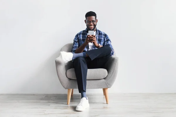 Guapo chico negro joven usando el teléfono celular, navegar por la web o las redes sociales, sentado en el sillón, comprobando la nueva aplicación móvil — Foto de Stock