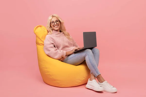 Ευτυχής freelancer κυρία χρησιμοποιώντας φορητό υπολογιστή, κάθεται σε καρέκλα beanbag και χαμογελώντας, τον έλεγχο της νέας ιστοσελίδας σε ροζ φόντο — Φωτογραφία Αρχείου