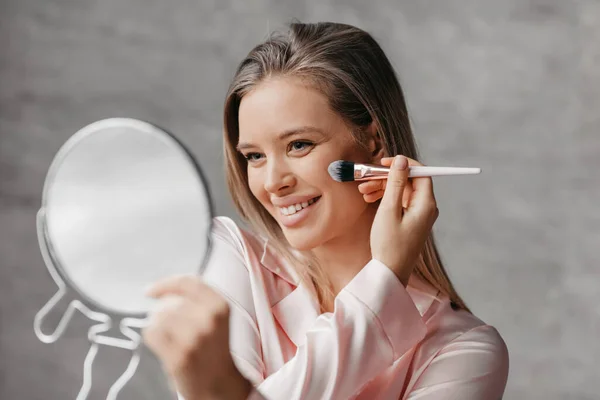 Ritual matinal para cuidados de beleza. Mulher bonita em sleepwear aplicando maquiagem no rosto com escova e olhando no espelho — Fotografia de Stock