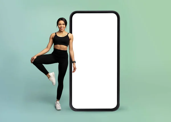 Çevrimiçi egzersiz için mobil uygulama. Genç fitness bayan büyük bir akıllı telefonun yanında duruyor beyaz beyaz ekranlı, model. — Stok fotoğraf