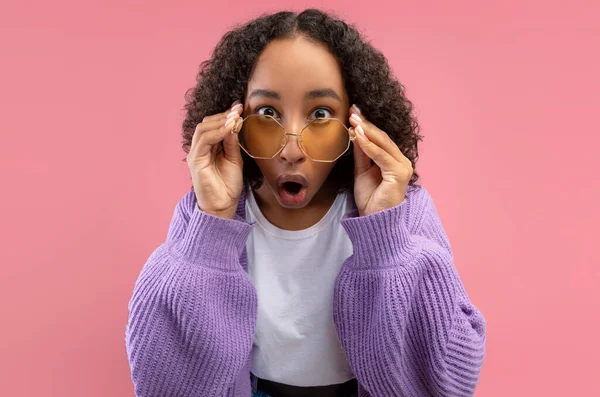 Millennial African American woman in trendy sunglasses looking at camera in shock on pink studio background — Stock Photo, Image