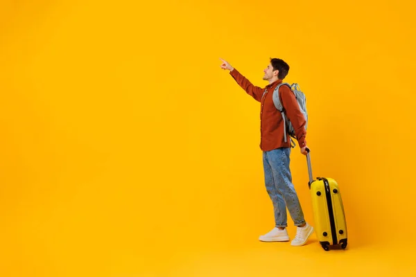 Turista hombre señalando dedo aparte de pie con la maleta, fondo amarillo — Foto de Stock