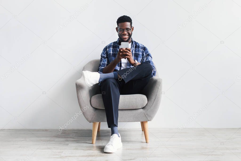 Handsome young black guy using cellphone, surfing web or social media, sitting in armchair, checking new mobile app