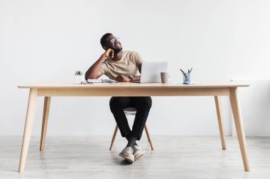 Masasında dizüstü bilgisayarla oturan mutlu siyah adam, hayal kuruyor, beyaz stüdyo duvarına karşı online çalışmaya ara veriyor.