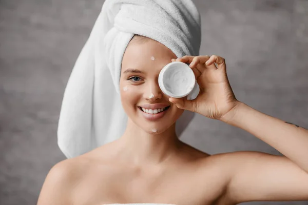 Nawilżająca skóra po prysznicu. Ładna milenijna kobieta w ręczniku zasłaniającym jej oko słoikiem śmietany we wnętrzu łazienki — Zdjęcie stockowe