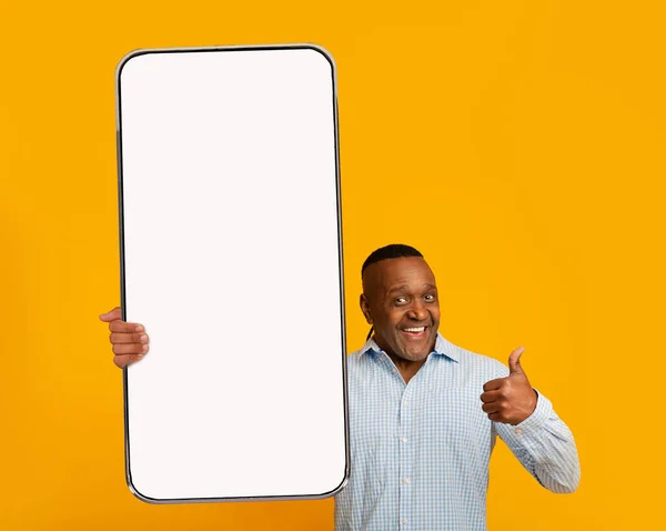Eu recomendo. maduro preto homem segurando enorme smartphone com branco branco tela na mão e mostrando polegar para cima, mockup — Fotografia de Stock